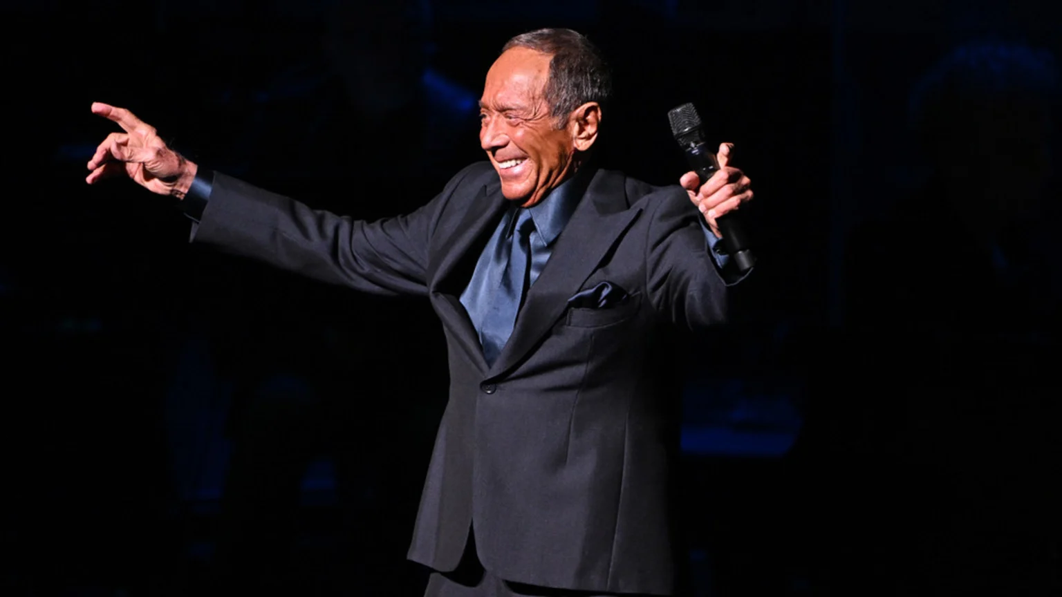 Paul Anka smiles broadly, holding a microphone and gesturing with his left arm while performing on a dark stage, dressed in a dark suit with a blue satin shirt and tie.