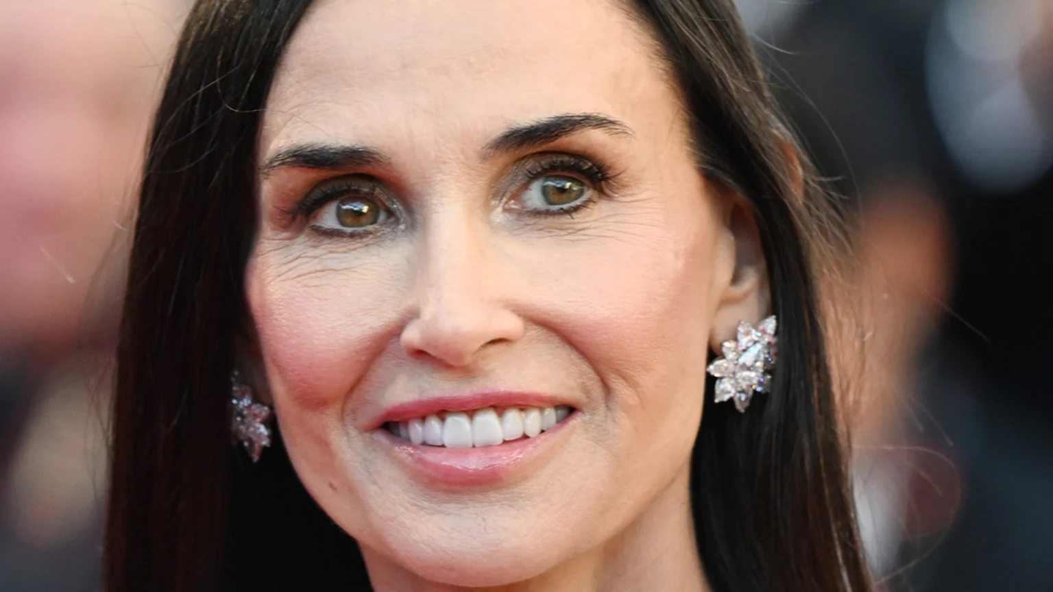 Close-up portrait of actress Demi Moore smiling, showing white teeth, wearing makeup and sparkling diamond earrings, with dark hair and a blurred background.