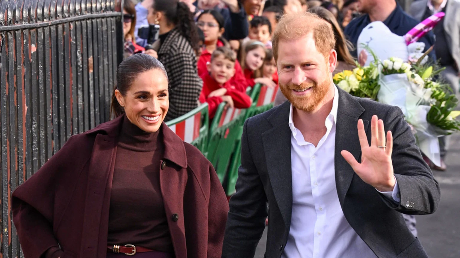 Meghan Markle in a burgundy coat and Prince Harry in a grey suit smile and wave to a crowd, including children, outdoors.