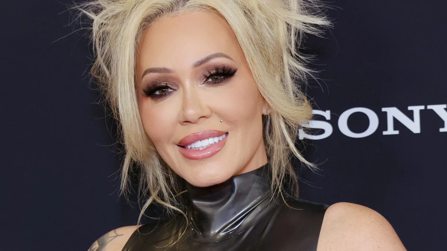 Close-up of a smiling woman with messy blonde hair, heavy eye makeup, full glossy lips, a facial piercing, and a shiny dark top, with a Sony logo behind her.