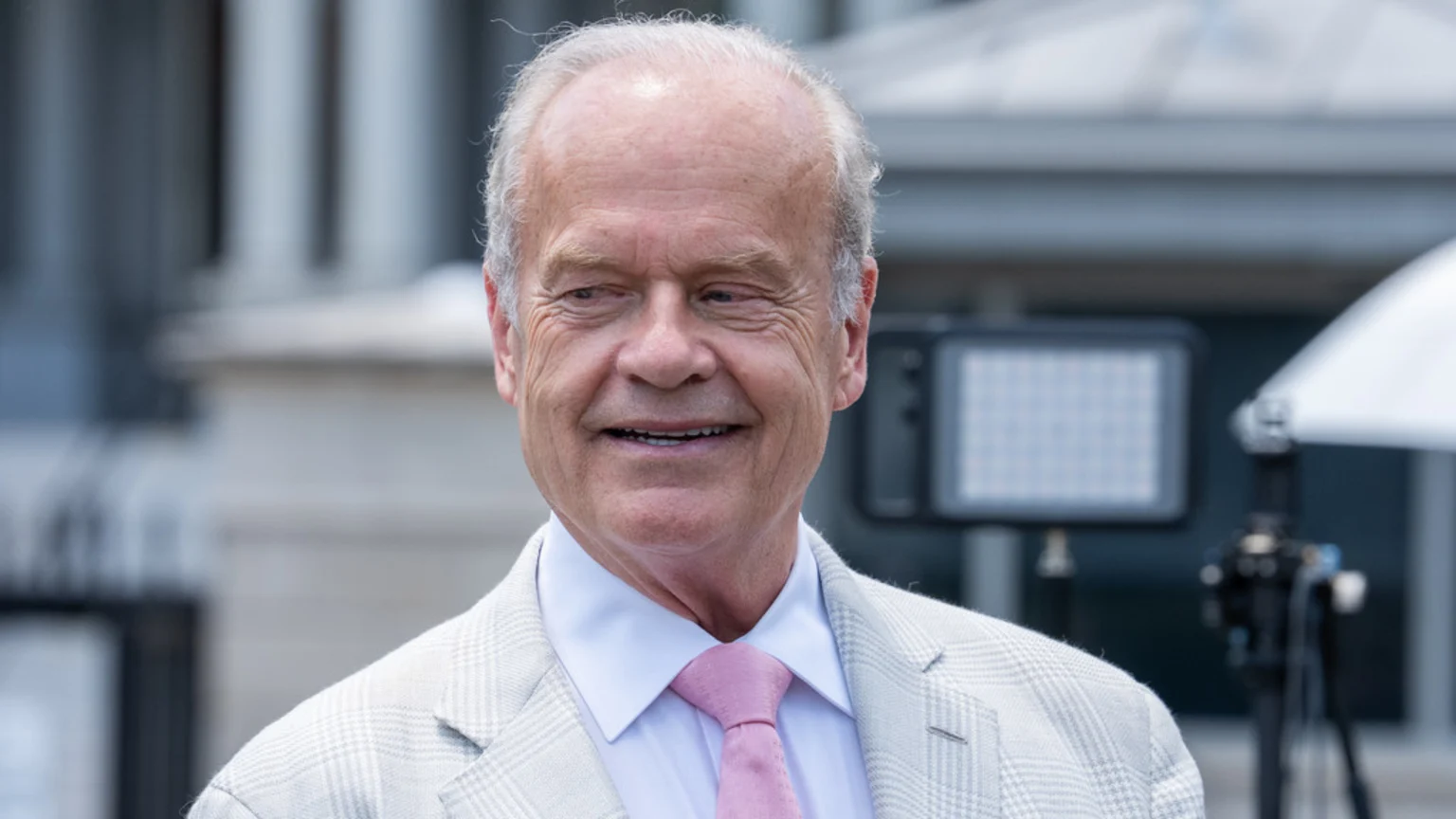 Kelsey Grammer, an older white man with gray hair, smiles slightly while wearing a light plaid suit jacket, white shirt, and pink tie outdoors.