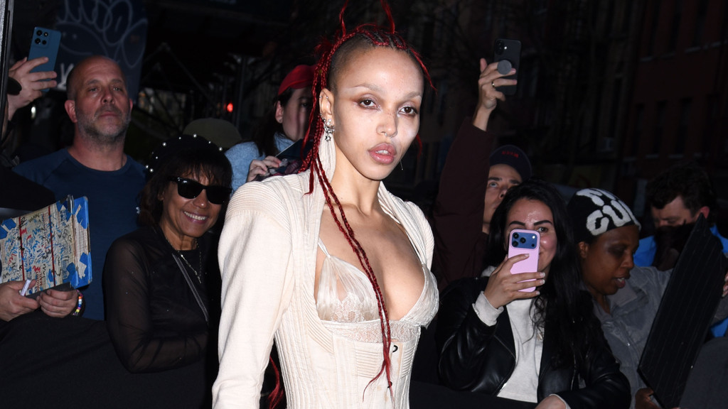 FKA Twigs, with red braids and unique makeup, poses for cameras while surrounded by a crowd of people holding up phones.