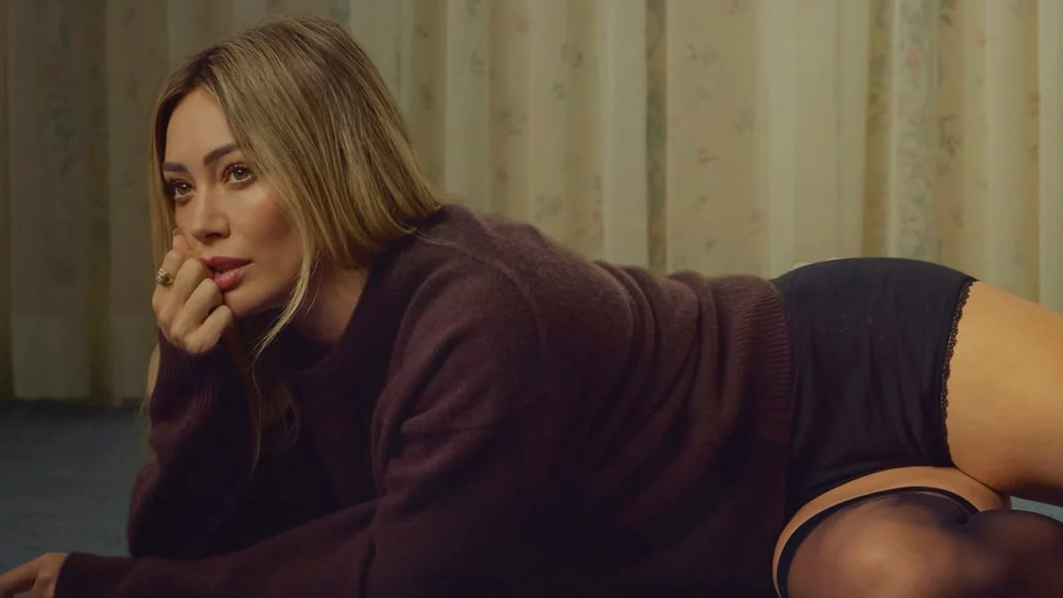 Hilary Duff lies on her stomach on a dark blue carpet, propped on her elbows with a thoughtful expression. She wears a dark brown sweater, black lace-trimmed high-waisted briefs, and sheer black thigh-high stockings. The background features light-colored curtains or wallpaper with a subtle floral pattern in a softly lit indoor setting.