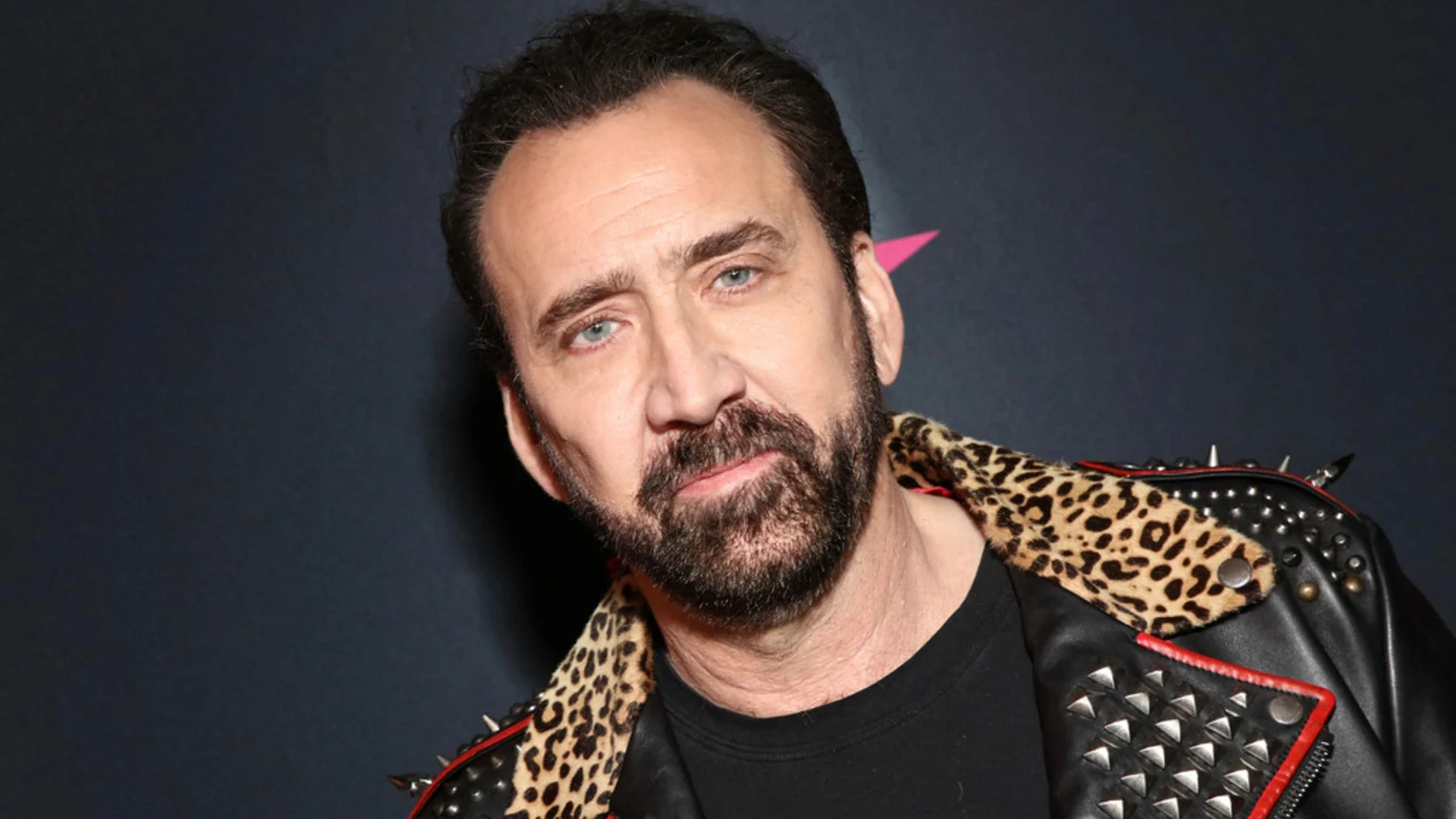 Nicolas Cage, with a beard and dark hair, wears a black studded leather jacket with a leopard print collar, looking at the camera.