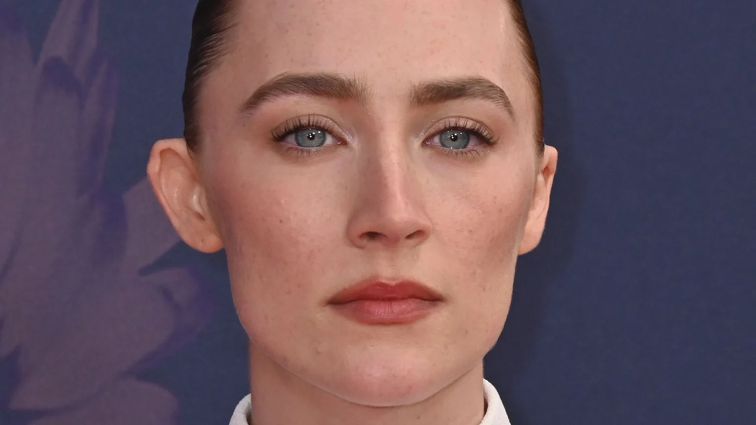 Close-up of Saoirse Ronan looking directly at the camera with a serious expression, her blue eyes, prominent eyebrows, and visible freckles framed by slicked-back hair against a dark background.