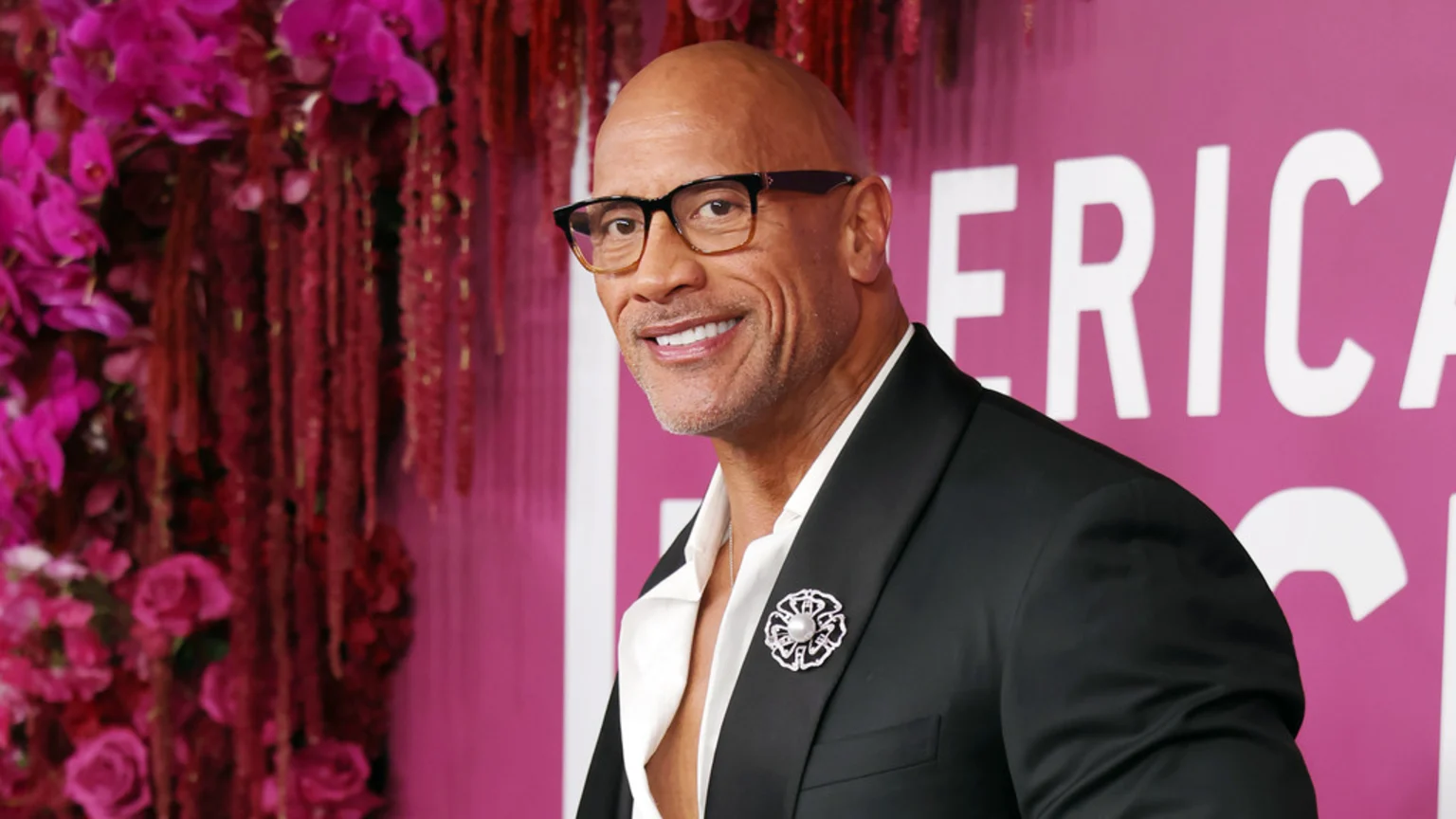 Dwayne Johnson smiles, wearing black glasses, a black suit, and a white shirt with a brooch, against a pink background with floral decor.