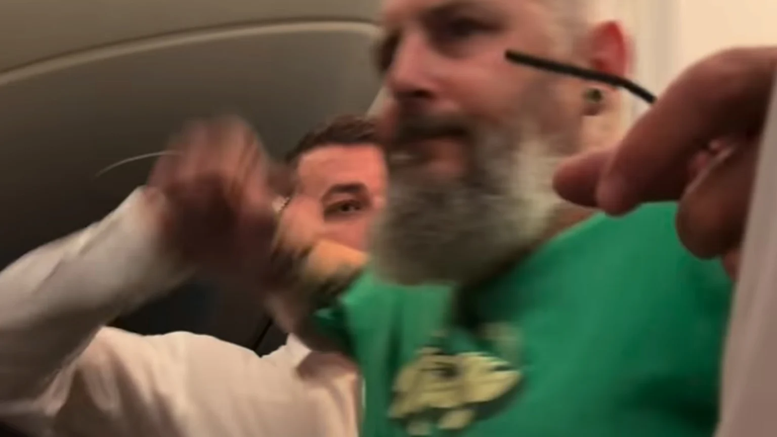 A blurred image captures a bearded man in a green shirt, with another man visible behind him, and multiple hands engaged in a dynamic interaction within a confined interior.