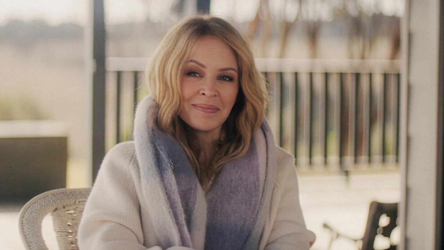 Kylie Minogue, with blonde wavy hair and a multi-colored fluffy scarf over a light sweater, smiles gently while sitting on a wicker chair in an outdoor setting.