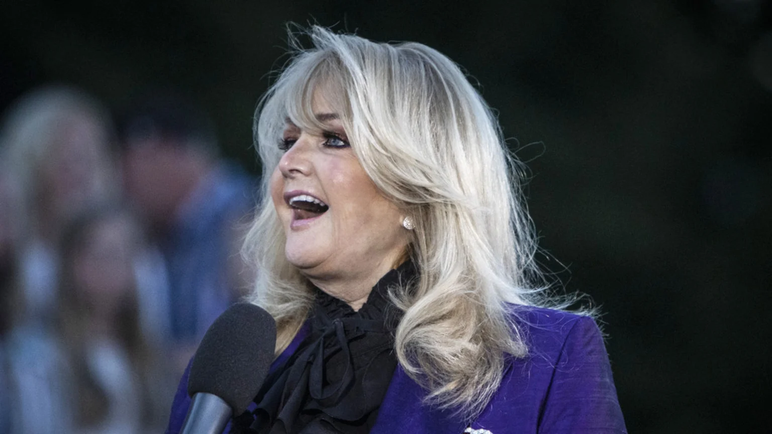 A blonde woman in a purple jacket and black ruffled blouse speaks into a microphone, smiling with her mouth open.