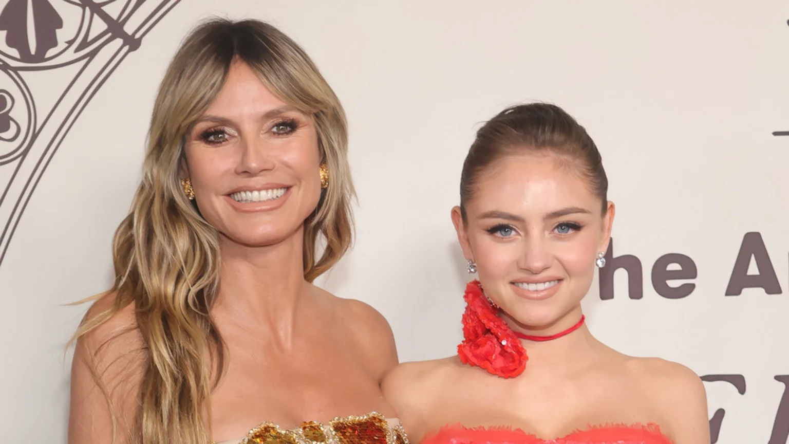 Heidi Klum, with long blonde hair and a gold sequined top, smiles next to her daughter Leni Klum, who wears a red strapless dress with a red neck piece.