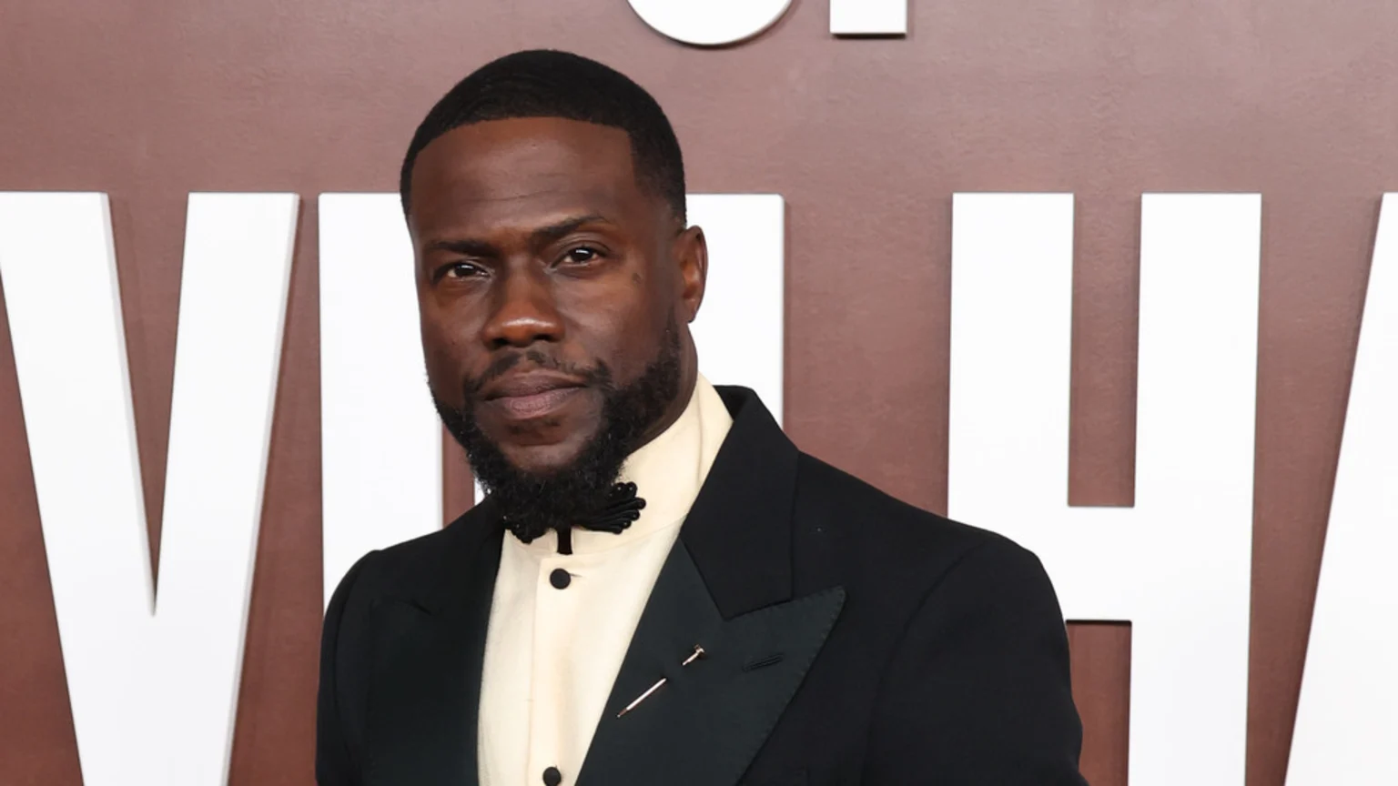 Kevin Hart, a Black man with a beard and short dark hair, wears a black suit, light shirt, a black decorative tie, and a lapel pin, looking forward against a brown background with white letters.