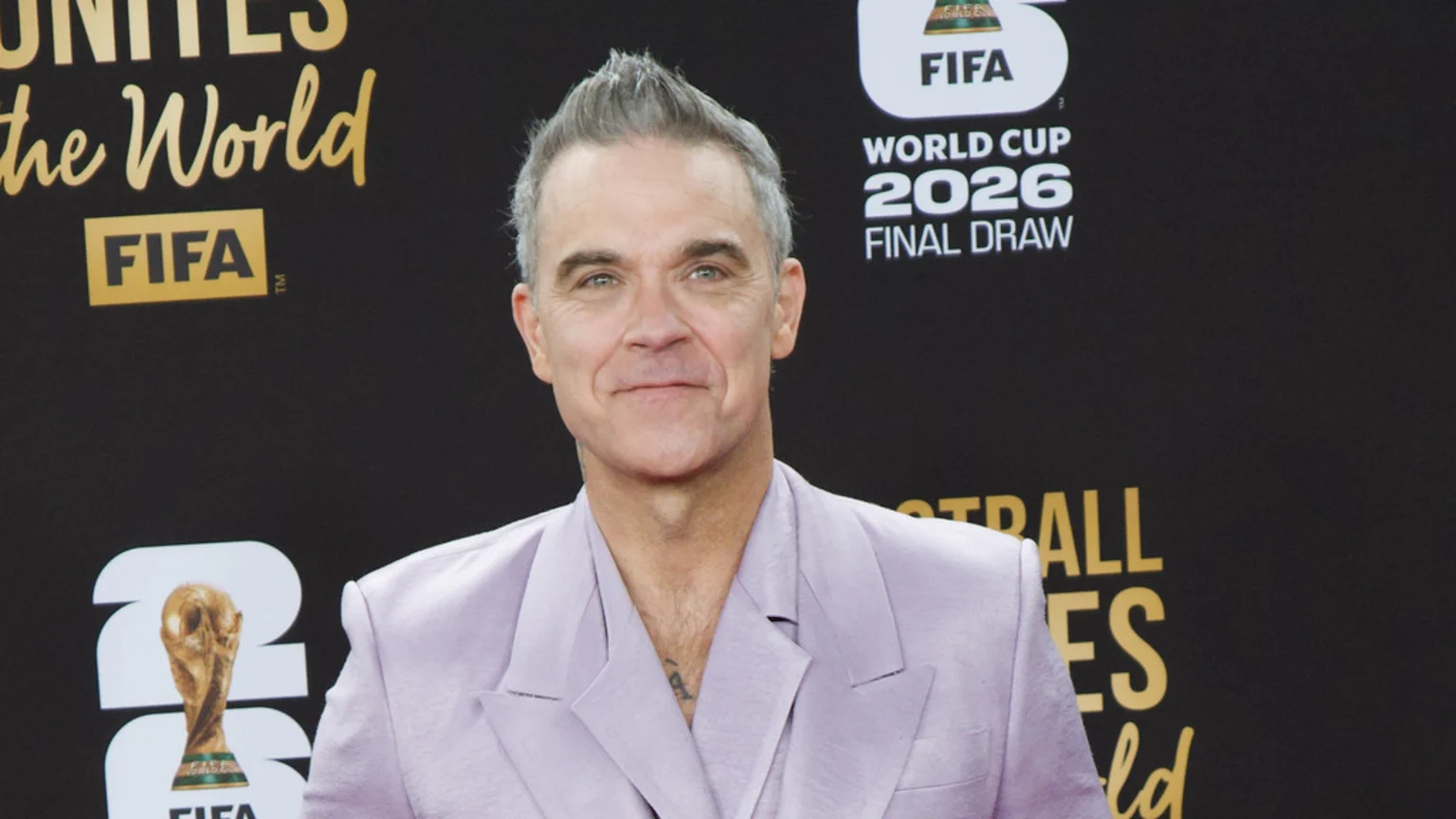 A man with grey, spiky hair and green eyes smiles slightly, wearing a light purple suit jacket, in front of a black background with FIFA and World Cup 2026 Final Draw logos.