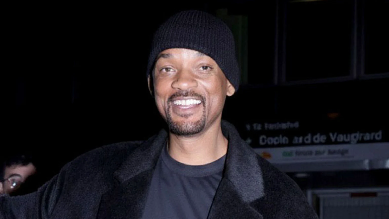 Actor Will Smith, wearing a black beanie and dark coat, smiles at the camera, with a dark, indistinct background showing some faint text.