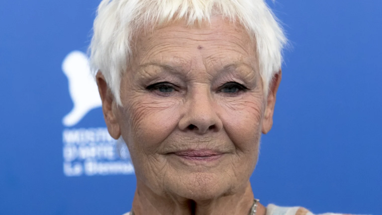 A close-up of an elderly woman with short white hair and blue eyes, smiling gently in front of a blue background with a blurred white logo.