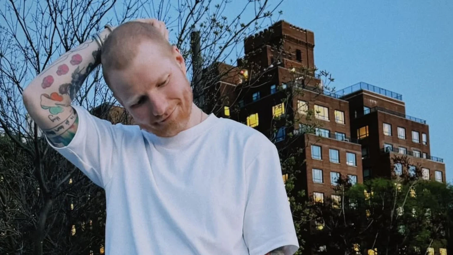 Ed Sheeran, smiling faintly, touches his head with his tattooed left arm, wearing a white t-shirt, with a multi-story brick building and tree branches visible in the background against a blue sky.