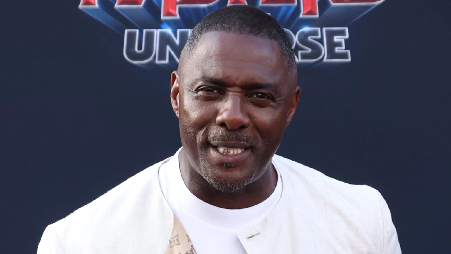 Actor Idris Elba smiles slightly at the camera, wearing a white t-shirt and a light-colored jacket, against a dark blue background with blurred text including "UNIVERSE".