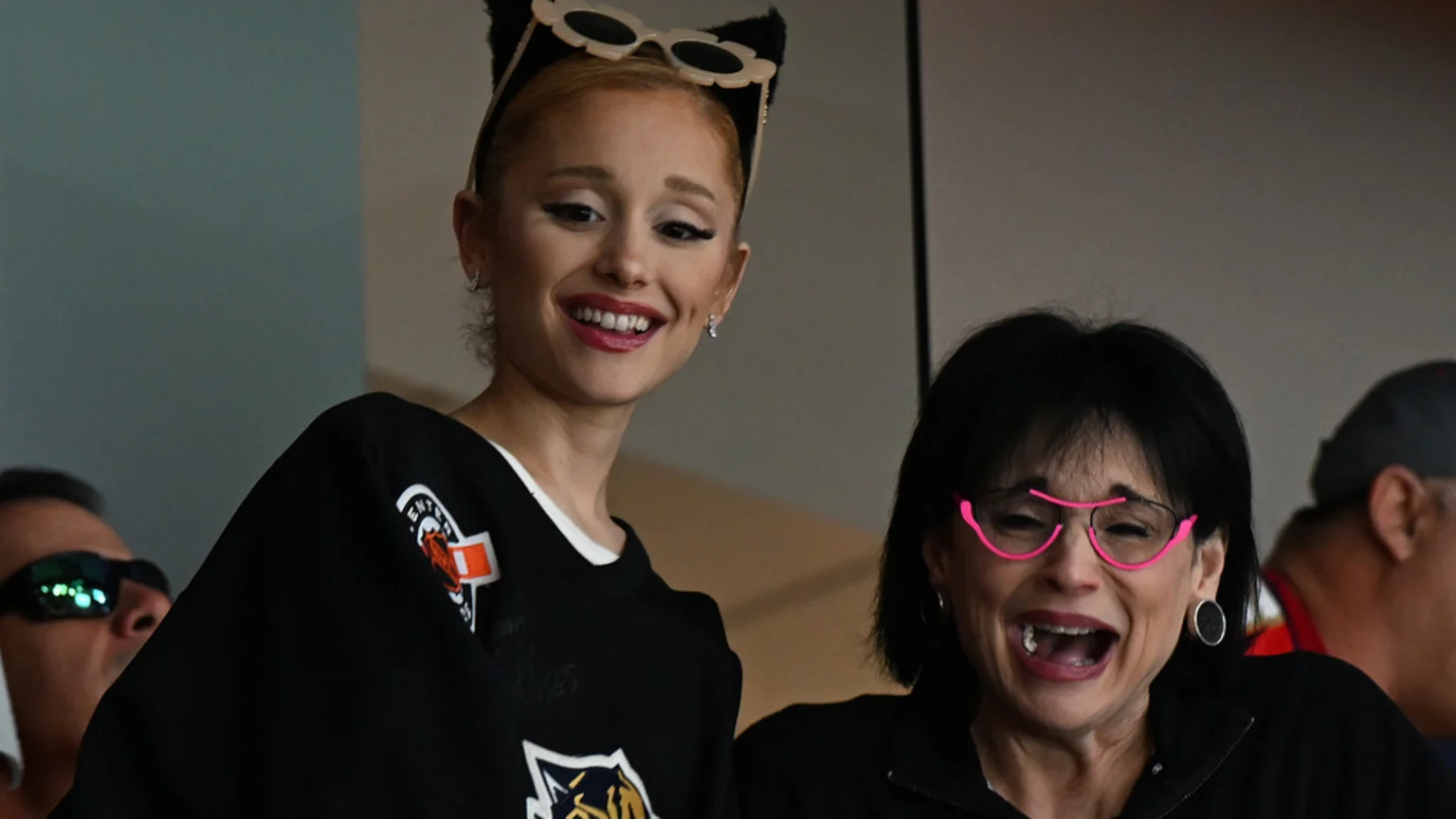 Ariana Grande, wearing cat-ear sunglasses and a black jersey, smiles next to a dark-haired woman in pink glasses who is laughing.