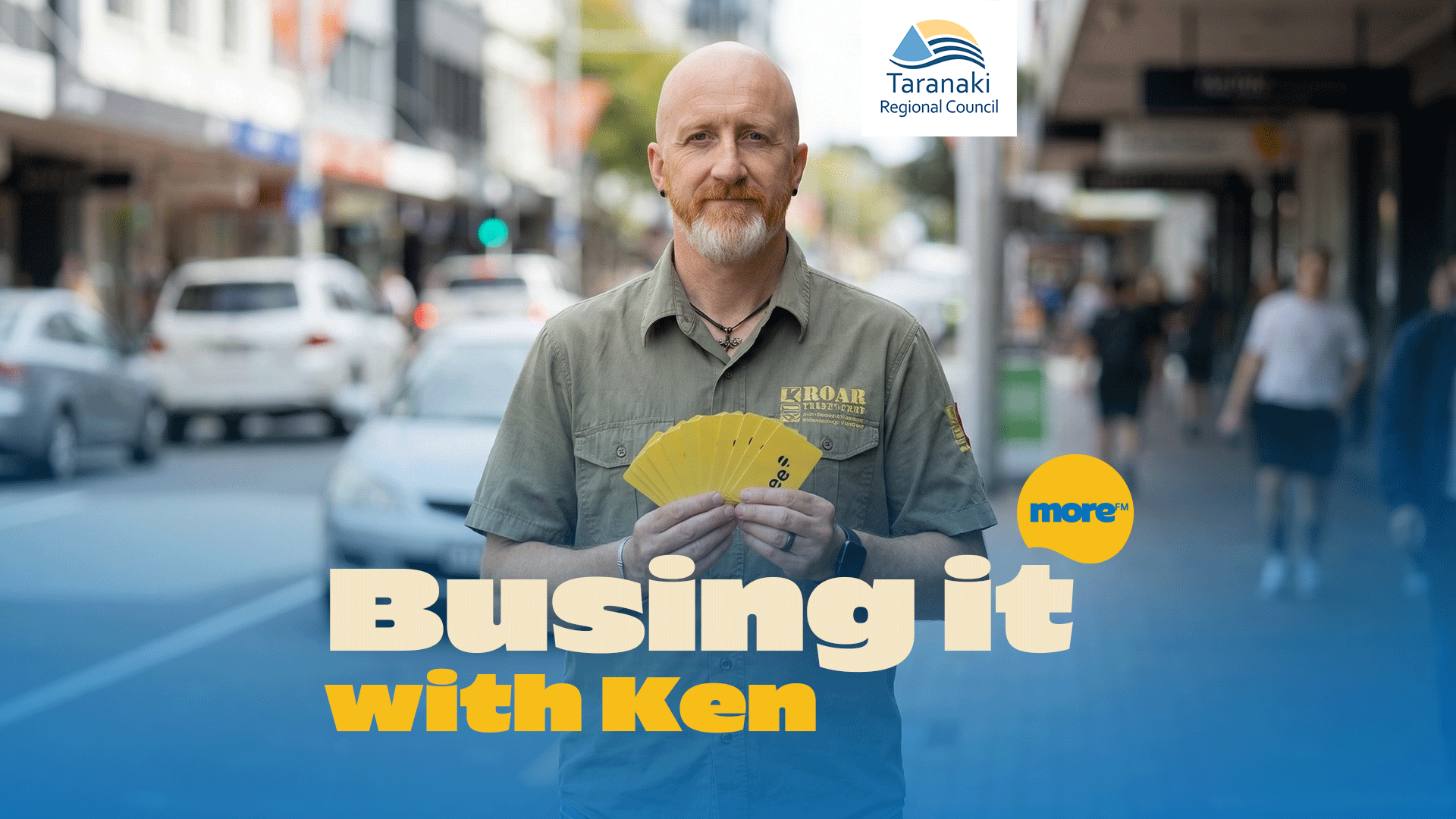 A bearded man holds yellow cards displaying "ees." in a city street, with "Busing it with Ken" text and Taranaki Regional Council and More FM logos.