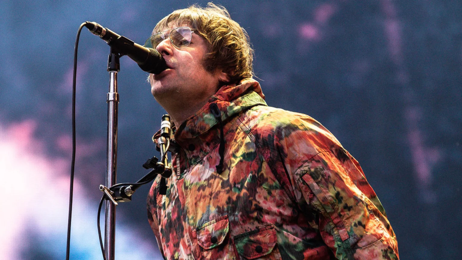 A man with light brown hair and sunglasses sings into a microphone, wearing a colorful floral camouflage jacket, against a dark, blurry background with pink and purple lights.