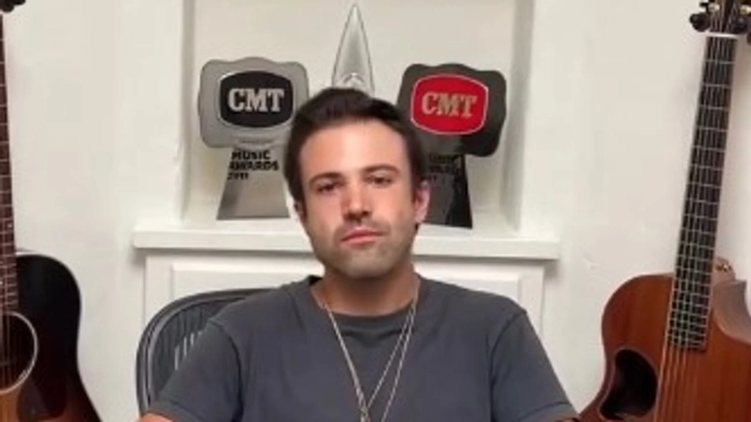 A man with dark hair and a grey shirt sits between two acoustic guitars, with two silver CMT Music Awards trophies and a clear trophy on a shelf behind him.