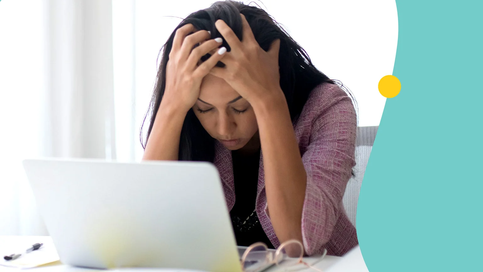 A woman with dark hair sits at a desk, looking stressed with her head in her hands, in front of a laptop, glasses, and a pen.