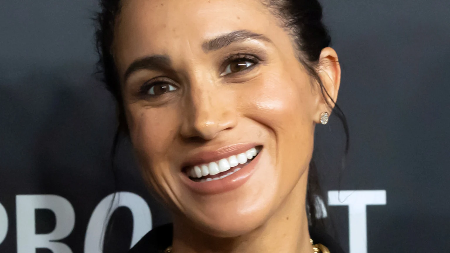 A close-up of Meghan Markle smiling brightly, showing her white teeth, with dark hair pulled back, wearing small stud earrings against a dark background with white lettering.