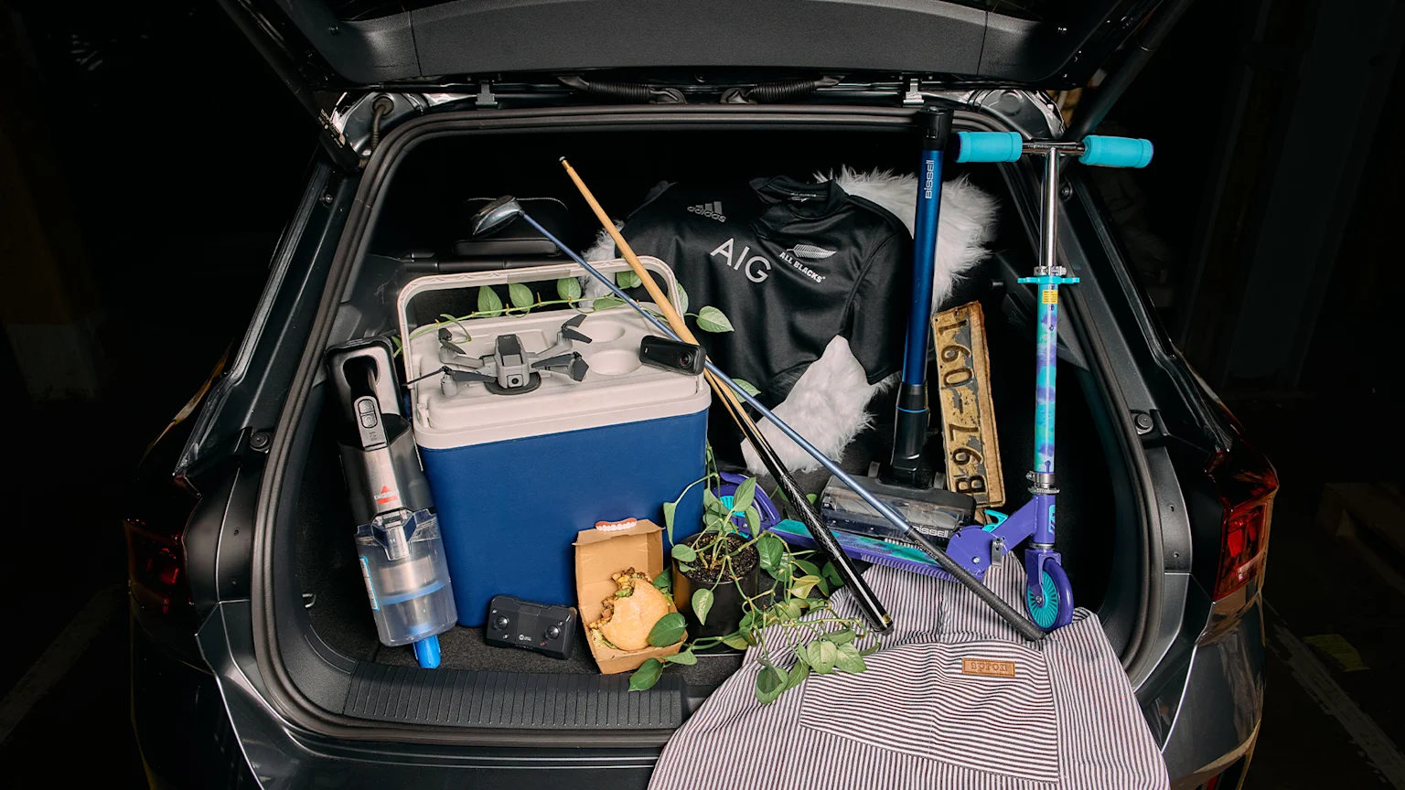 An open car trunk is packed with diverse items including a cooler, a drone, a rugby jersey, a Bissell vacuum, a scooter, a golf club, a pool cue, a license plate, a potted plant, a burger, and false teeth.