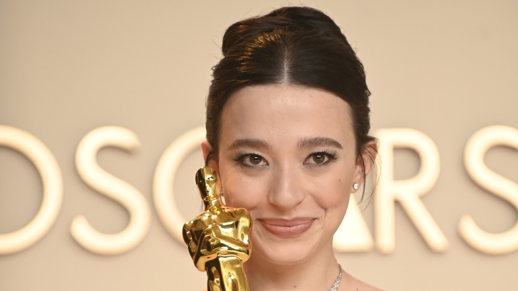 A woman with dark hair pulled back smiles faintly, holding a golden Oscar statuette in front of a blurred "OSCARS" sign.