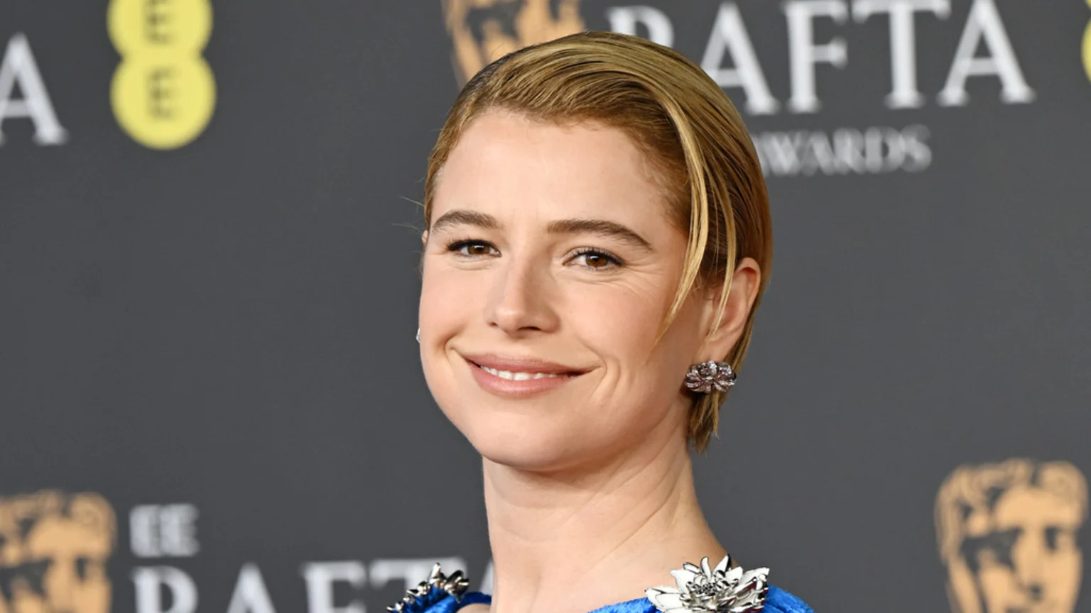 A smiling woman with short blonde hair and a blue embellished dress looks at the camera against a blurry dark background with "BAFTA" and "EE" logos.