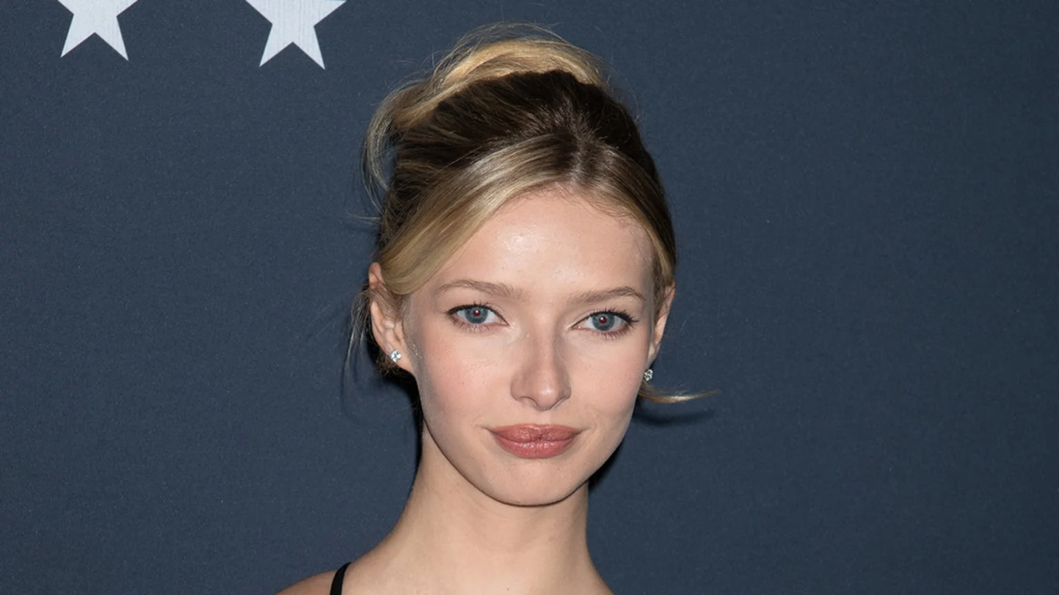 A studio-lit headshot of a young woman with blonde hair in a high bun and diamond stud earrings. She poses with a neutral expression and slight smile against a dark grey indoor event backdrop featuring two white five-pointed stars.