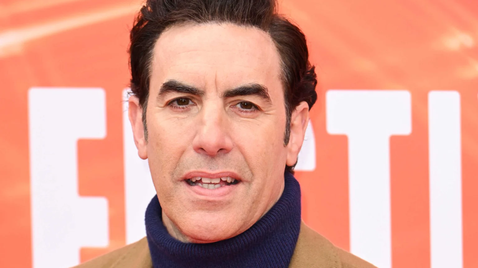 Close-up of a man with dark hair, prominent eyebrows, and brown eyes, wearing a blue turtleneck and a tan jacket, smiling slightly in front of an out-of-focus orange background with white letters.