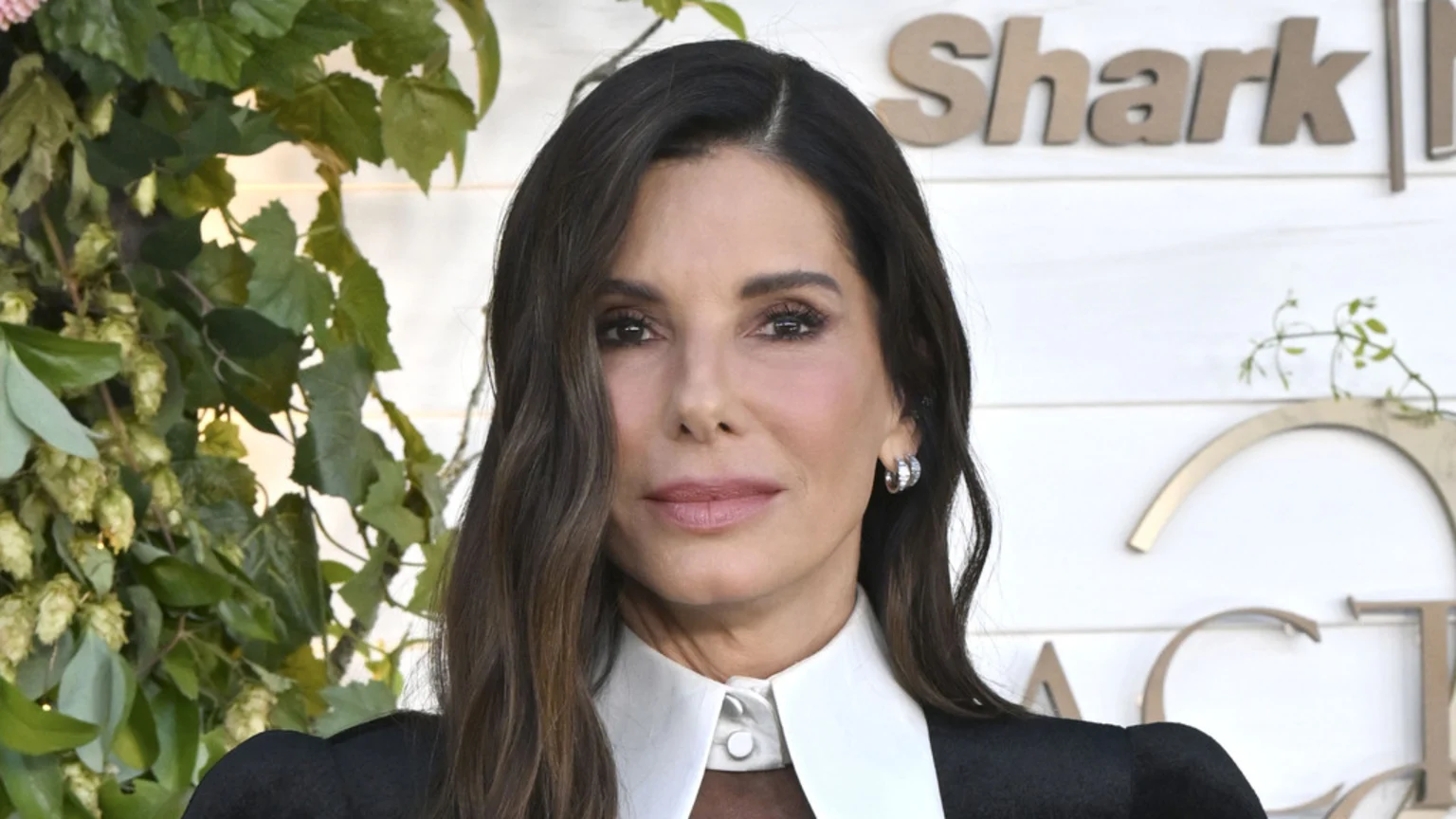 Actress Sandra Bullock with long dark hair and a white collared shirt looks at the camera, with green foliage on the left and "Shark" wooden letters on a white wall on the right.
