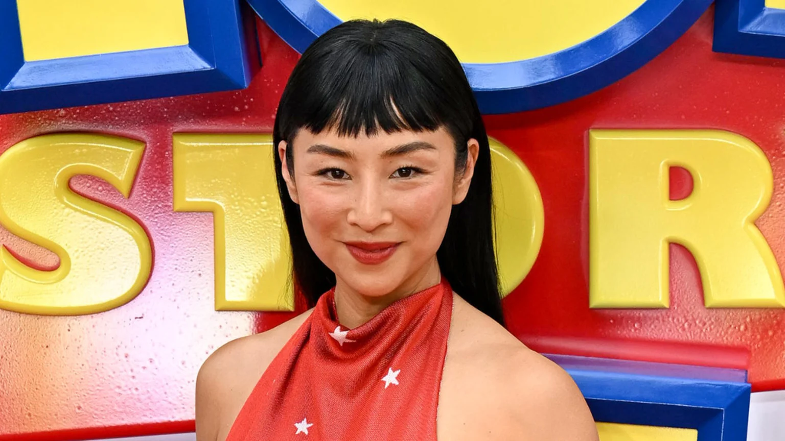 Greta Lee, with dark hair and bangs, smiles softly in a red top with white stars, against a colorful "Toy Story" background.