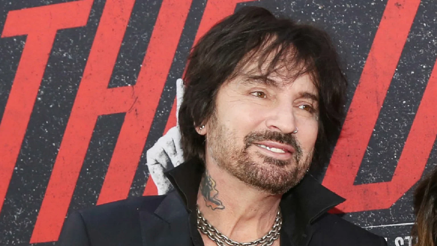 A man with dark messy hair, a beard, nose ring, and a neck tattoo smiles, wearing a black collared shirt and chain necklace, against a textured red and black background.