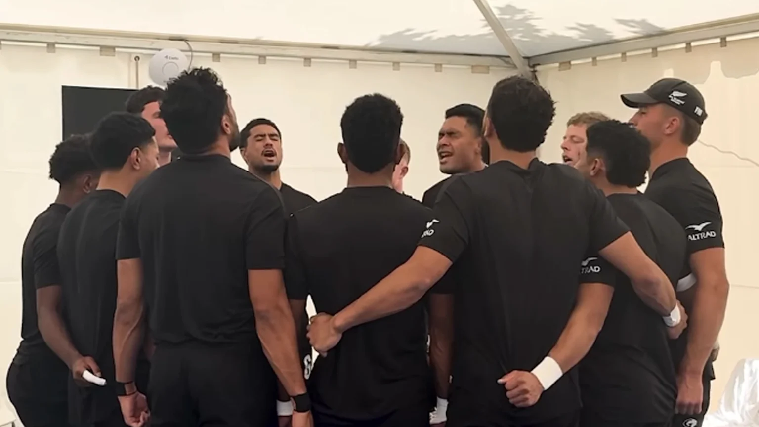 A group of All Blacks Sevens rugby players in black shirts stands in a huddle, singing or chanting with arms around each other in a pre-game tradition.