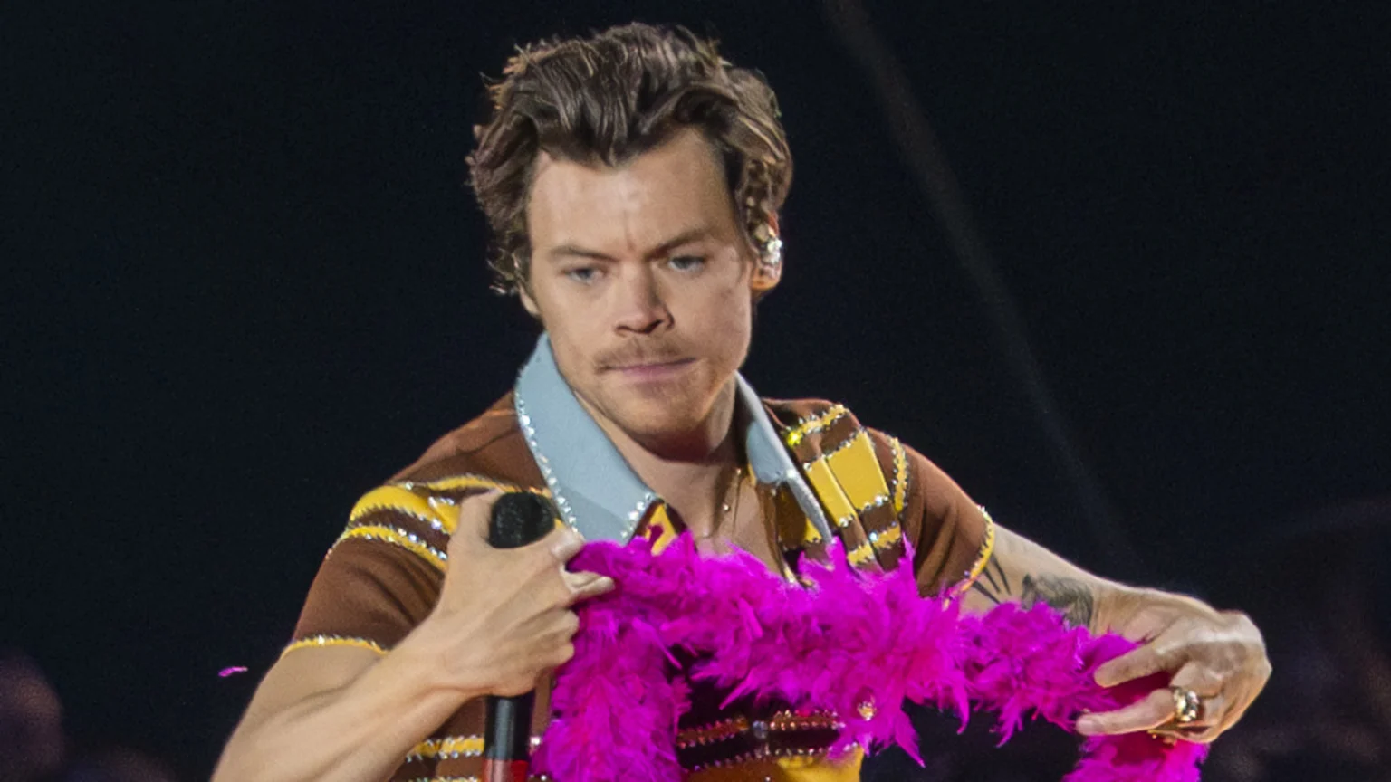 Harry Styles on stage, wearing an embellished brown and yellow striped shirt with a light blue collar, holding a microphone and a bright pink feather boa.