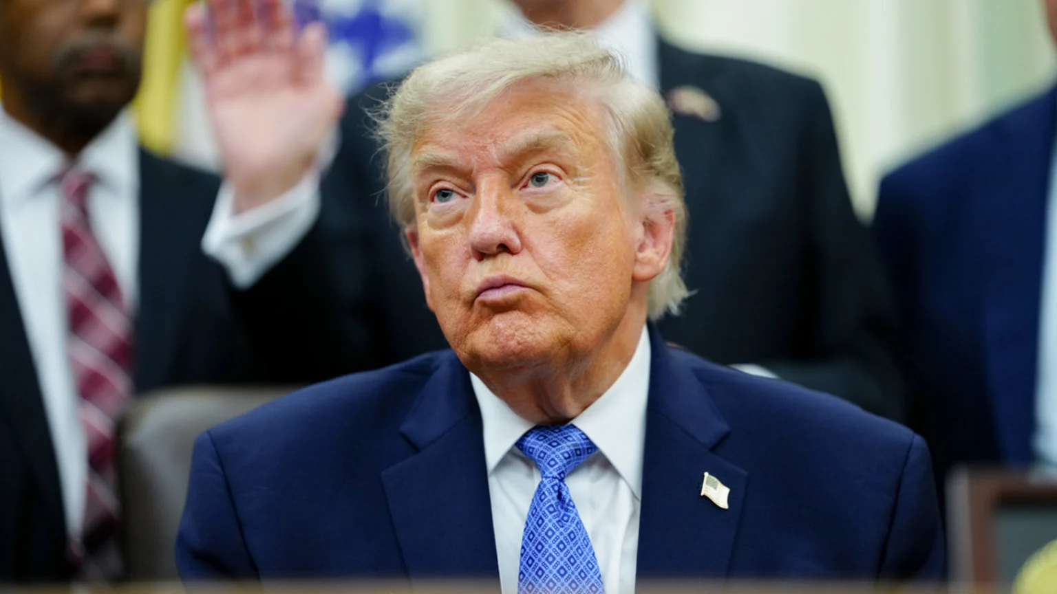 Close-up of Donald Trump looking pensive, wearing a dark blue suit, blue patterned tie, and an American flag lapel pin, with blurred figures in the background.