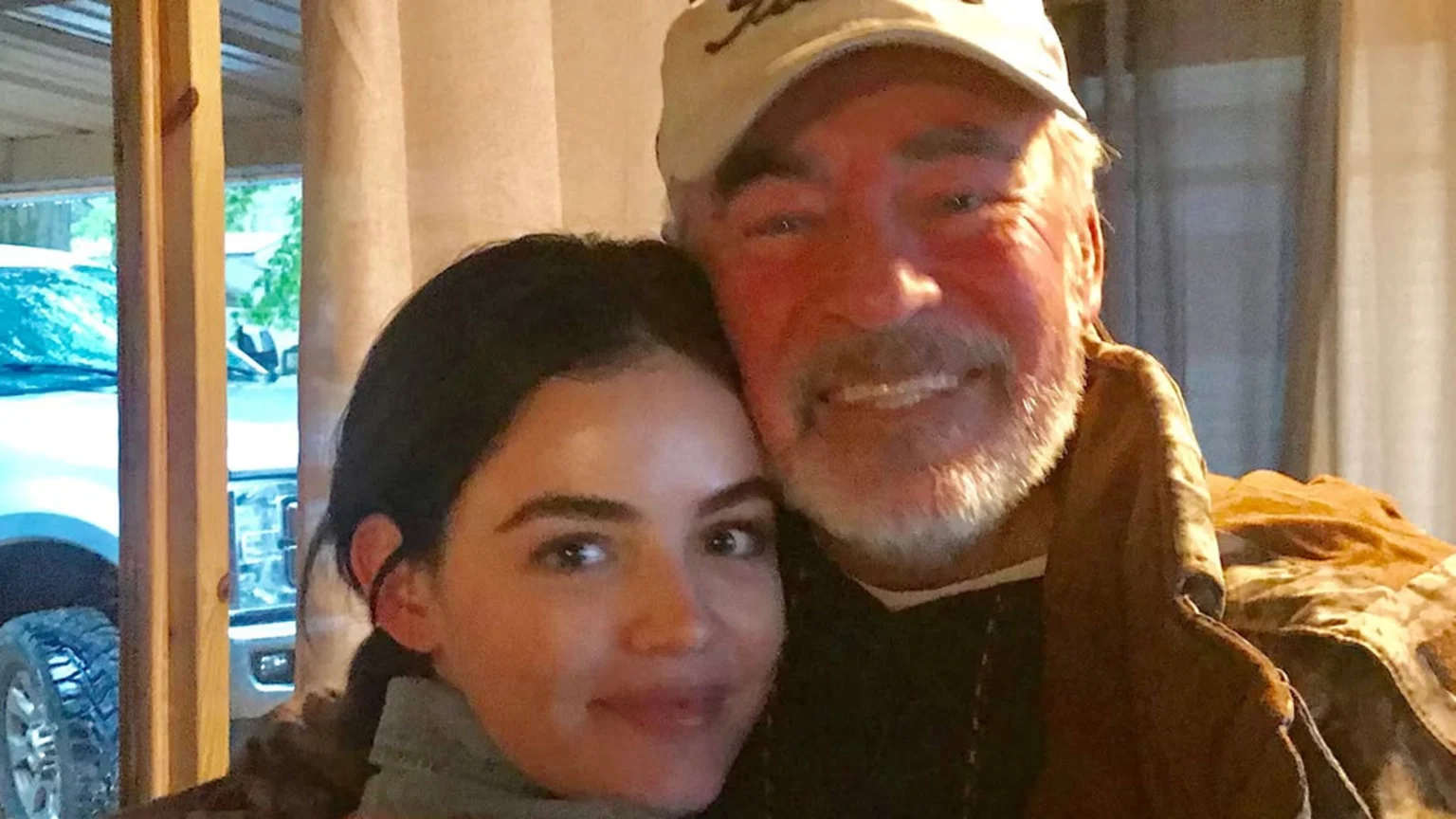 A young woman and an older man with a grey beard and baseball cap embrace and smile warmly for a close-up photo indoors.
