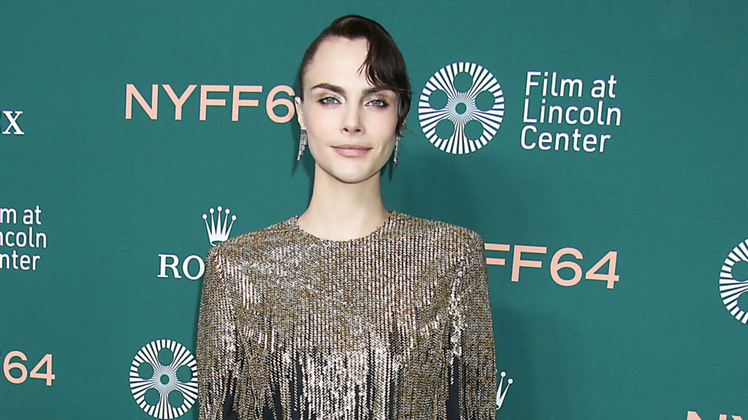 Cara Delevingne poses in a sparkly golden fringe top against a teal backdrop with "NYFF64", "Film at Lincoln Center", and "Rolex" logos.