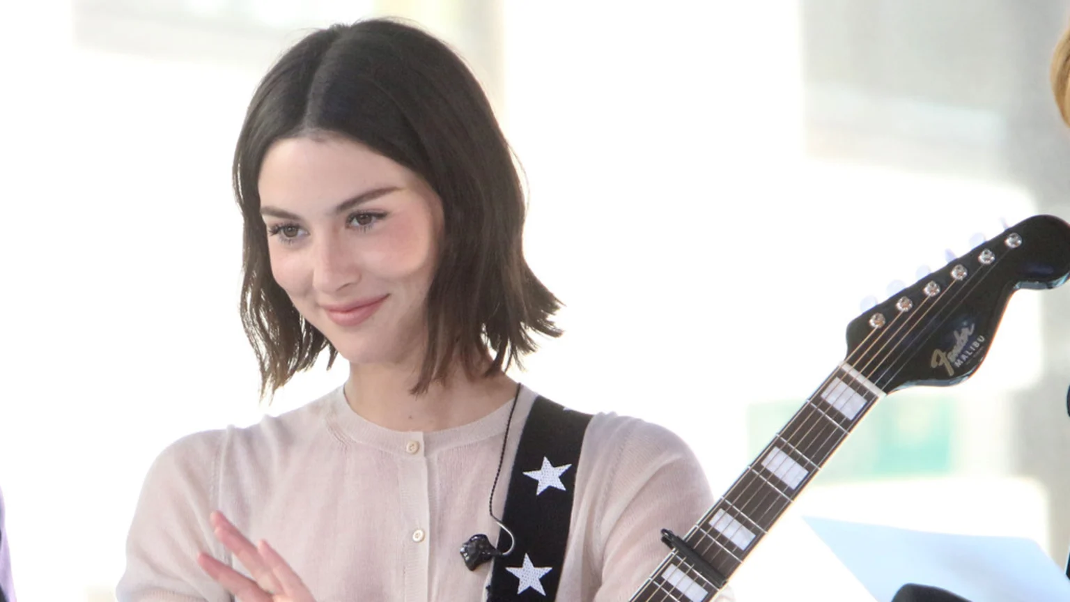 Gracie Abrams, smiling slightly with dark hair, wears a light cardigan and a star-patterned guitar strap with an in-ear monitor, holding a Fender Malibu acoustic guitar.