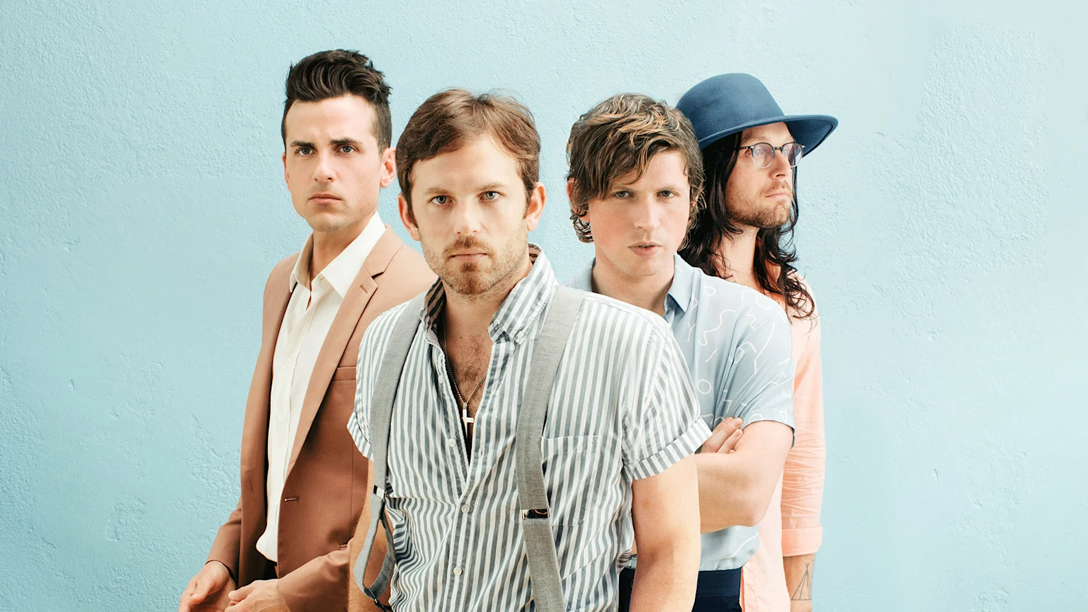 Four serious-looking men stand against a light blue wall: one in a brown suit, one in a striped shirt and suspenders, one in a light blue patterned shirt, and one in a blue hat with glasses and a peach shirt.