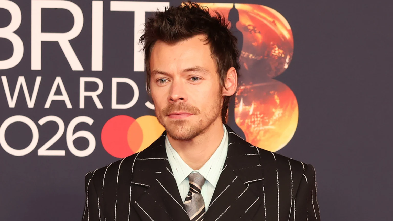 Harry Styles wearing a dark pinstripe suit, light striped shirt, and striped tie, against a backdrop with "BRIT AWARDS 2026", the Mastercard logo, and an orange Brit Award trophy.