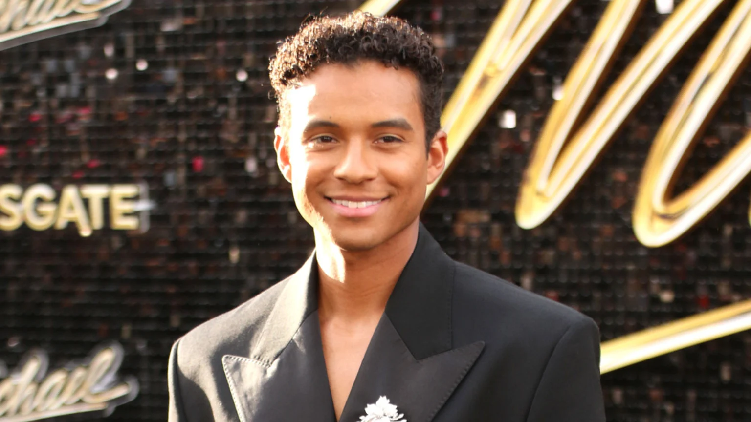 A smiling young man with curly dark hair wears a black suit jacket adorned with a sparkly brooch, standing against a blurry dark background featuring golden text.