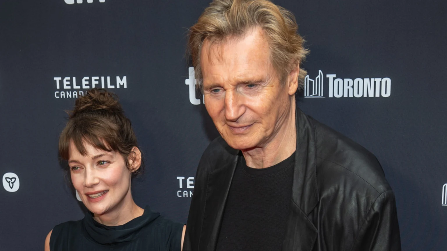 Liam Neeson, an older man in a black leather jacket, stands next to Stella Stocker, a woman with brown hair in a bun, against a dark backdrop with event logos.
