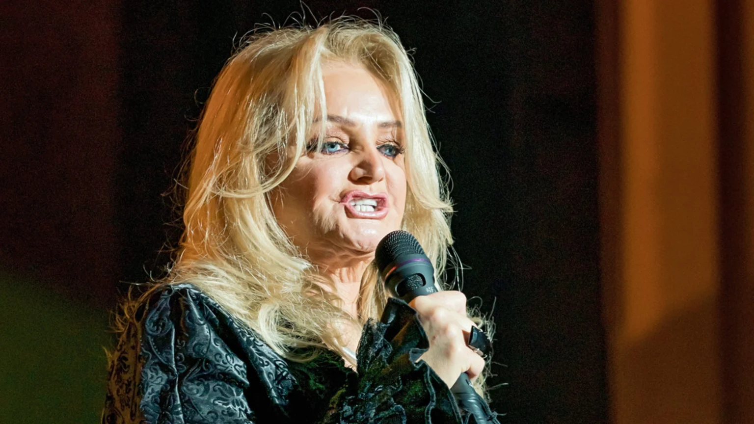 A woman with long blonde hair, blue eyes, and makeup sings into a black microphone, wearing a dark patterned top against a dark, blurred background.