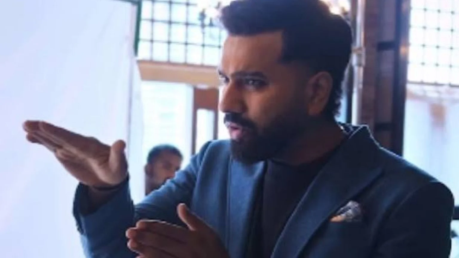 A man with styled hair and a beard, wearing a blue blazer, gestures with both hands, looking right, against a background with a window.
