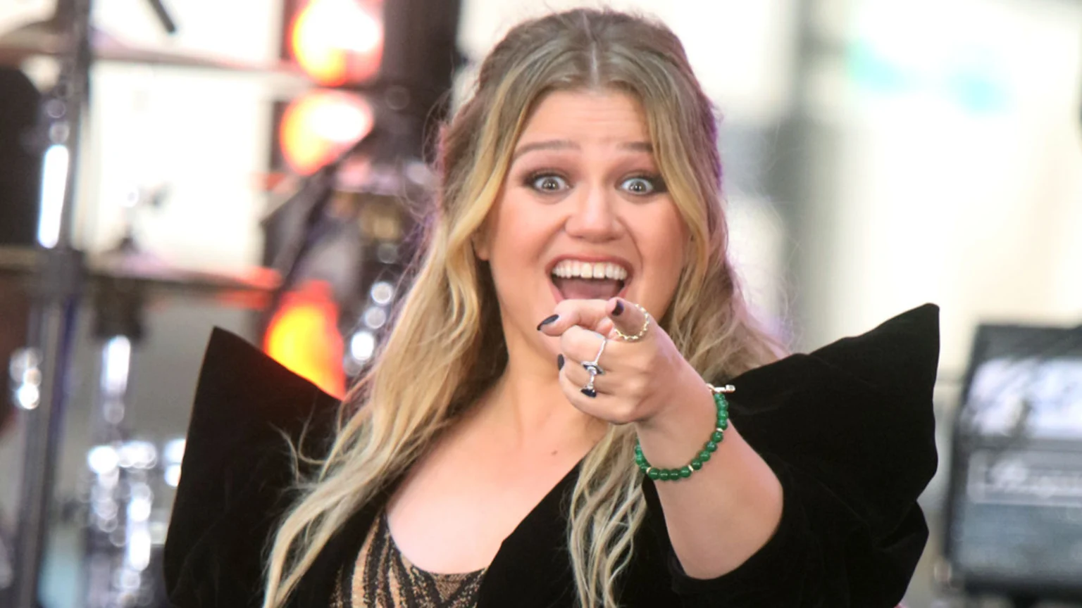 Close-up eye-level portrait of Kelly Clarkson smiling widely and pointing forward on a brightly lit stage. She wears a black structured top with patterned garment underneath, adorned with rings and a green beaded bracelet. Background shows blurred warm-toned stage lights and metallic equipment.