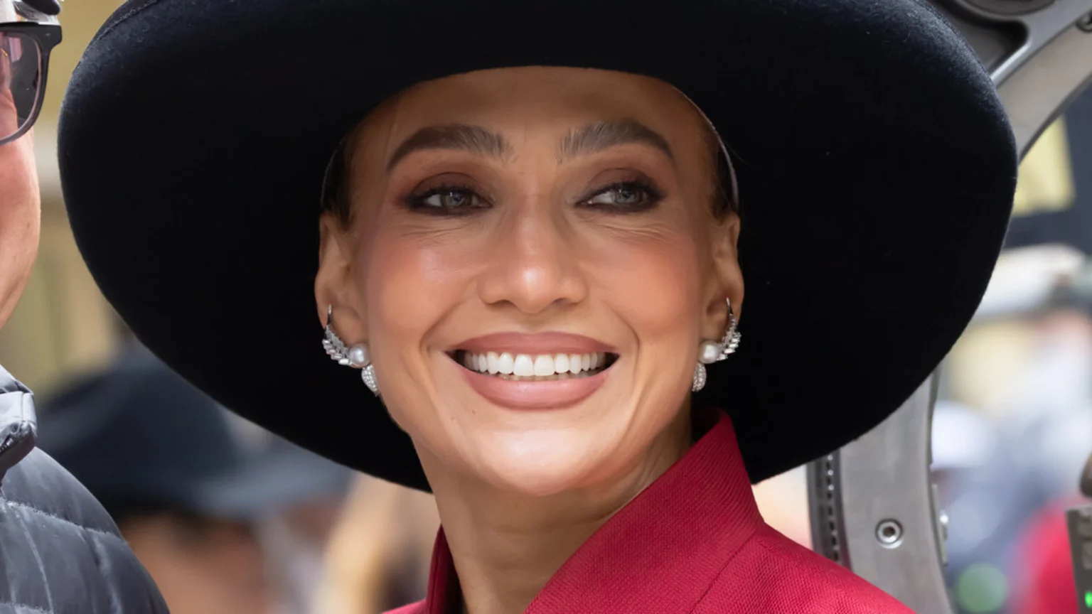 Jennifer Lopez smiles broadly, wearing a black hat, a red top, and ornate pearl and diamond earrings.