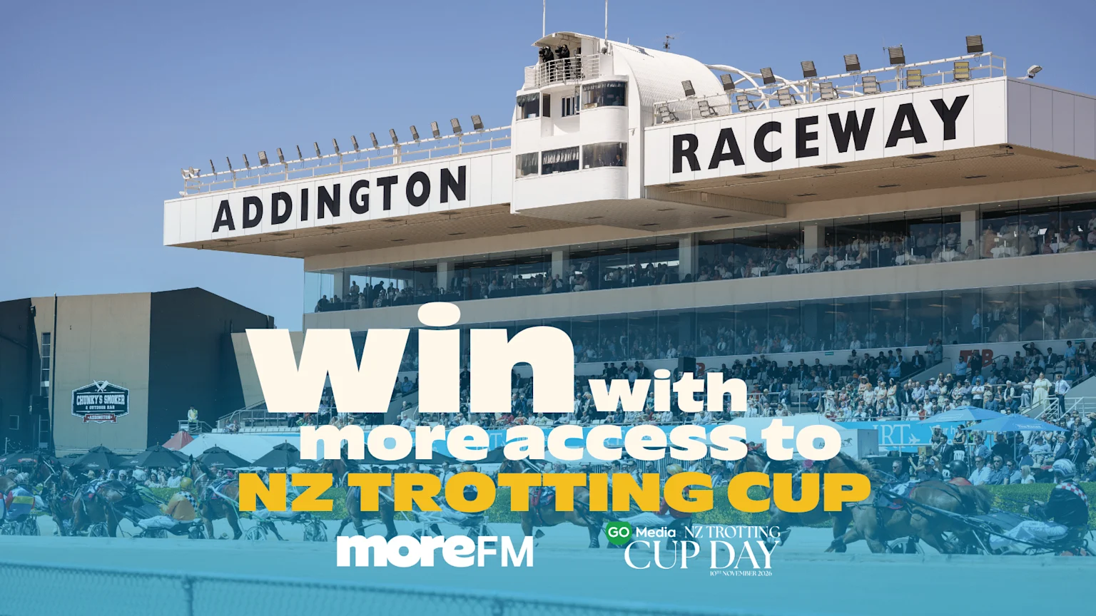 A harness race in progress at Addington Raceway with spectators in the stands, overlaid with promotional text reading "Win with more access to NZ TROTTING CUP" from MoreFM for "GO Media NZ TROTTING CUP DAY 10th November 2026."