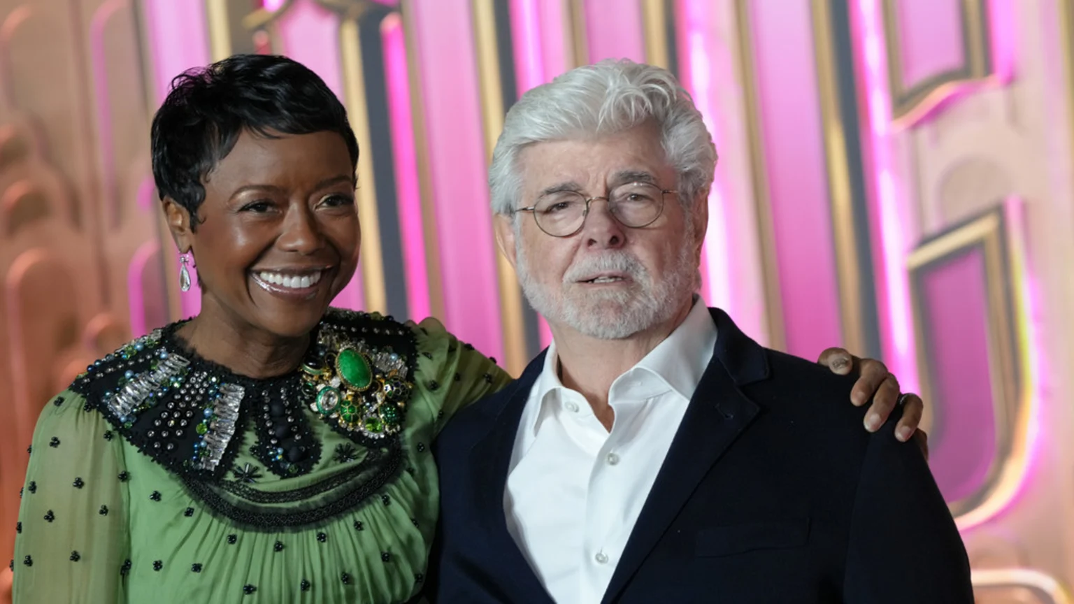 George Lucas, a man with white hair and beard, stands with a smiling woman in a green beaded dress with an ornate collar and large brooch, her hand on his shoulder, against a blurred pink background.